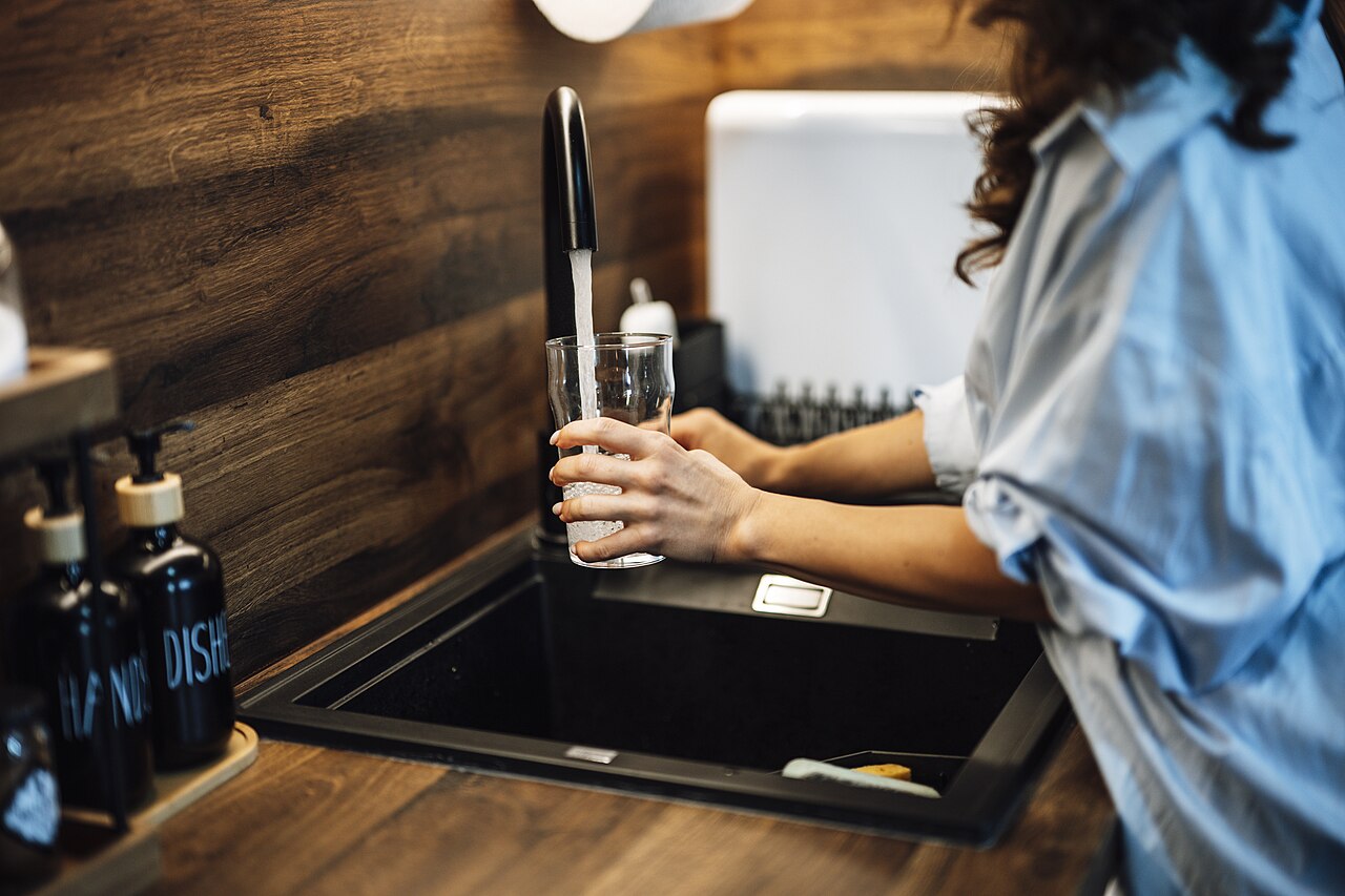 A person filling a clear glass from a kitchen faucet