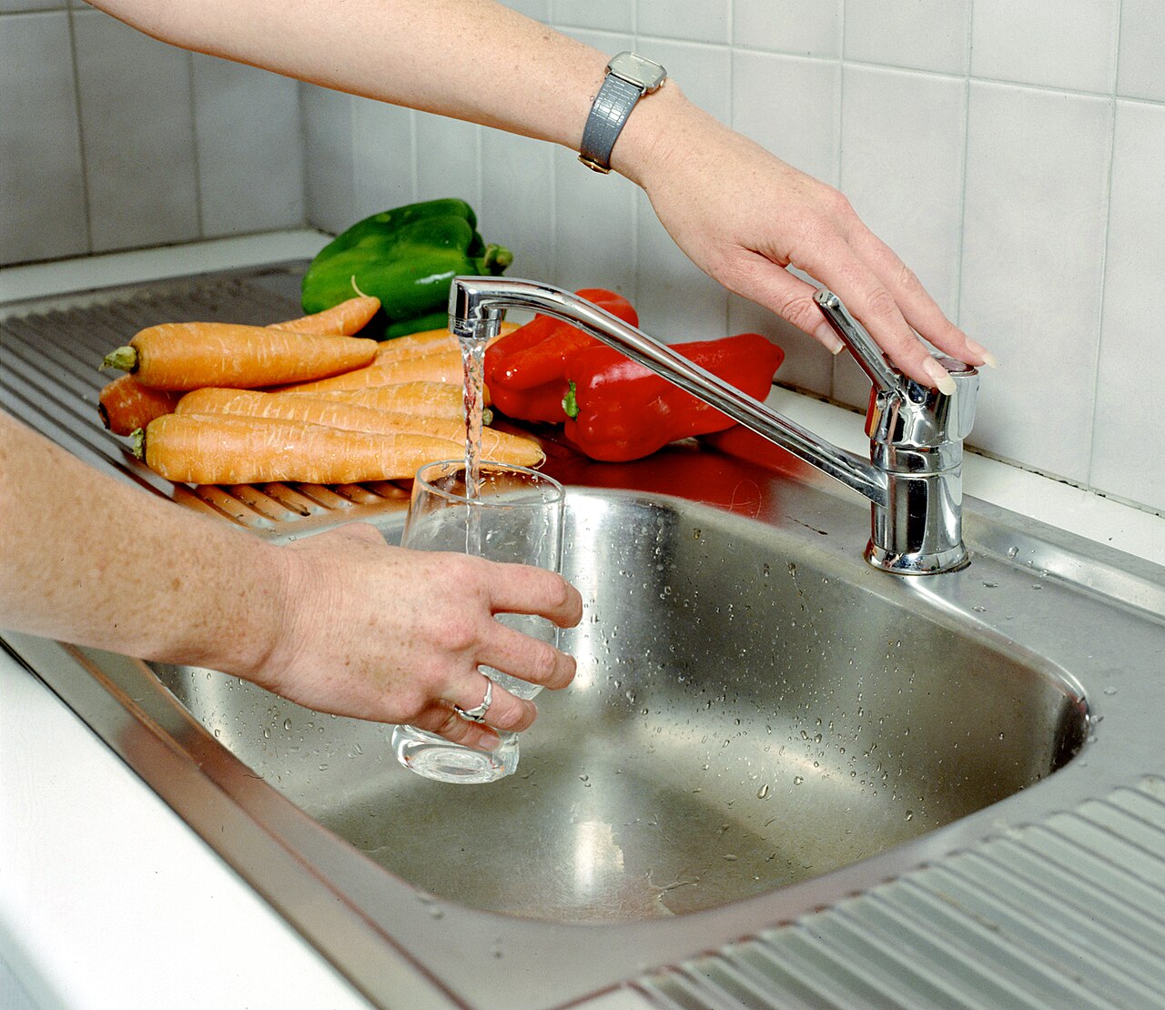 A person filling a glass from a kitchen tap beside fresh vegetables
